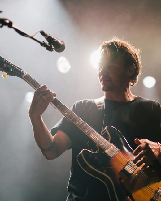 A guitarist mid-performance under a hard white spotlight, backlit by haze.
