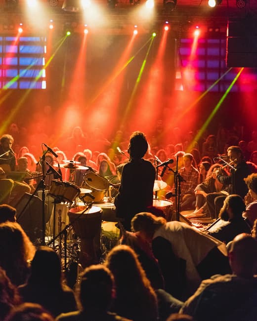 A drummer seen from behind the kit, playing to an audience under red and pink stage lighting.