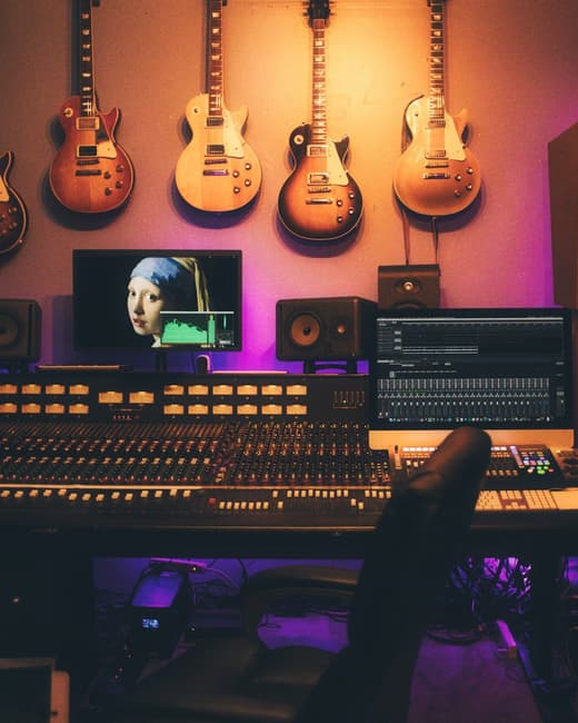 A recording studio control room with a large mixing console and electric guitars racked on the wall behind it.