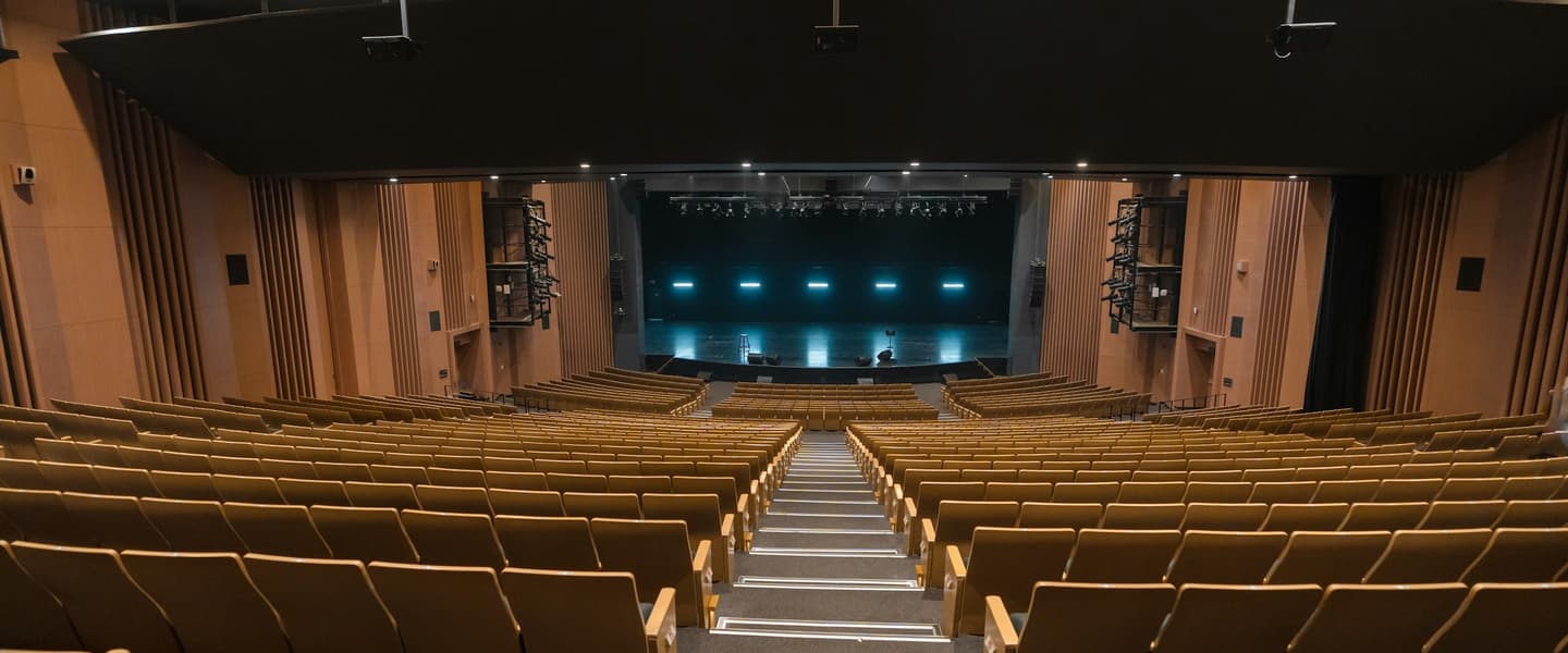 An empty auditorium of wooden seats facing a lit but bare stage.