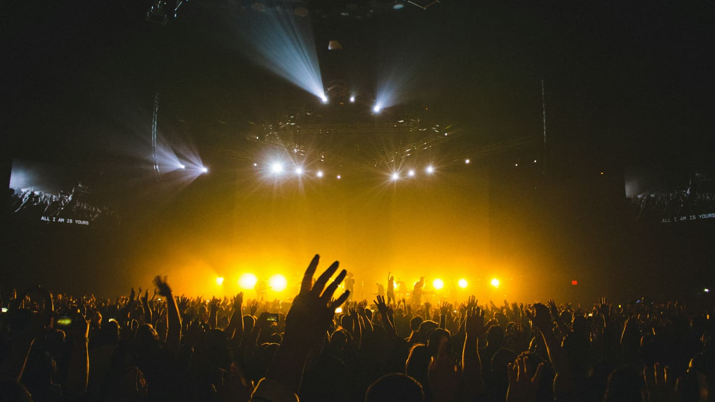 A crowd in silhouette in front of a wall of warm stage lights at a live show.