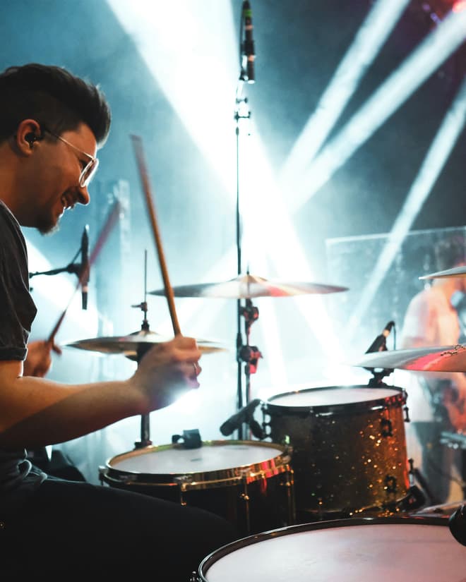 A drummer mid-strike behind a kit, lit from behind by hard white beams.