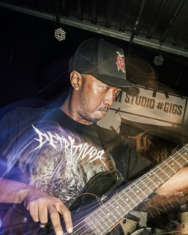 A bass player in a cap and dark shirt playing under blue stage lighting.